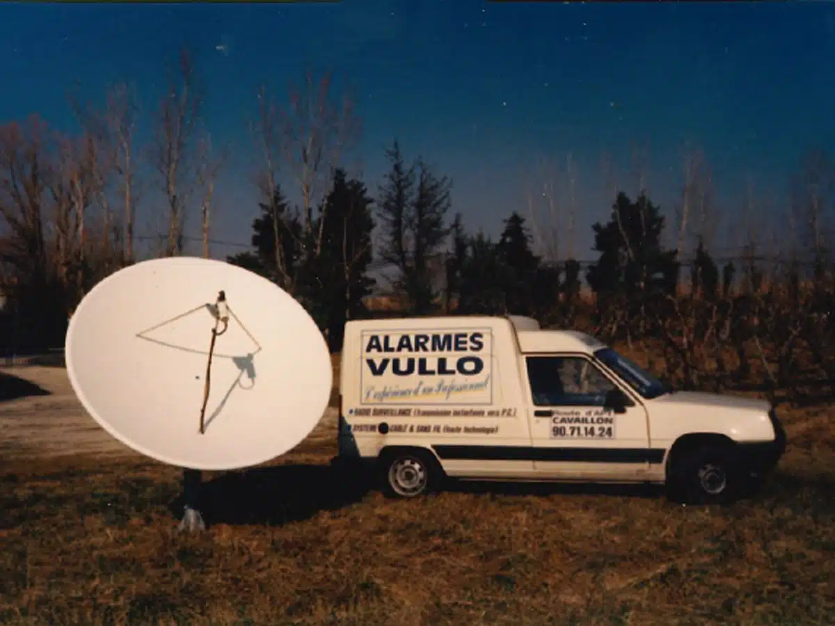 Vieille voiture avec alarme Vullo sous un ciel étoilé près d’une parabole géante.